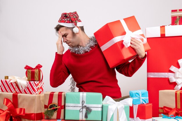 Vue de face confus jeune homme assis autour de cadeaux de Noël