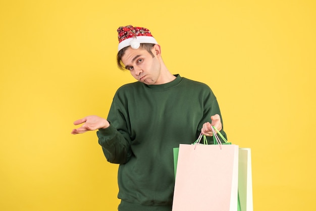 Vue de face confus jeune homme avec Bonnet de Noel tenant des sacs à provisions debout sur jaune