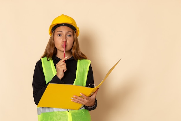 Vue de face constructeur féminin en casque jaune tenant un document jaune et un crayon sur un mur blanc