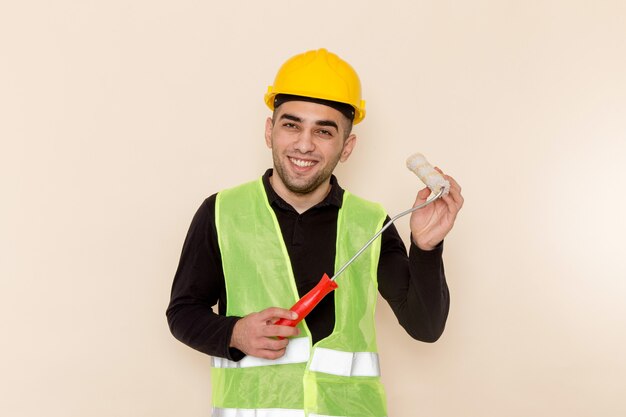 Vue de face constructeur masculin en casque jaune tenant le rouleau et souriant sur le bureau crème