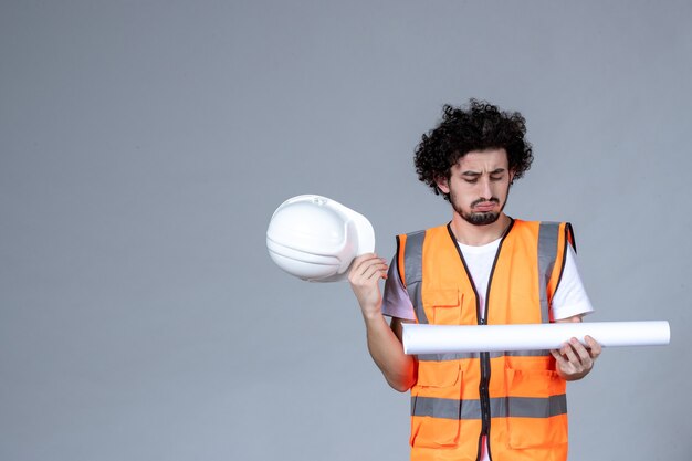 Vue de face d'un constructeur masculin concentré dans un gilet d'avertissement tenant un casque de sécurité et montrant un blanc sur un mur gris