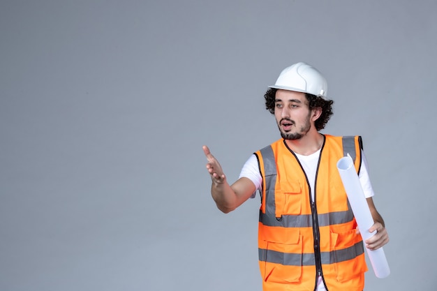 Vue de face d'un constructeur masculin concerné dans un gilet d'avertissement portant un casque de sécurité et tenant un blanc pointant vers l'avant sur un mur gris vague