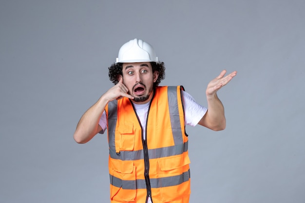 Vue de face d'un constructeur masculin confus portant un gilet d'avertissement avec un casque de sécurité et faisant un geste d'appel sur le mur de la vague grise