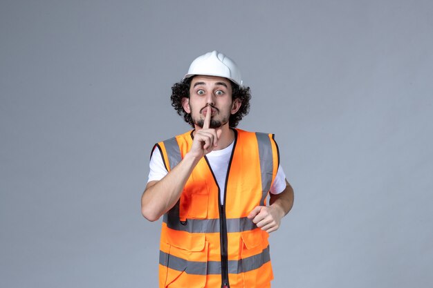 Vue de face d'un constructeur masculin nerveux portant un gilet d'avertissement avec un casque de sécurité et faisant un geste de silence sur le mur de la vague grise