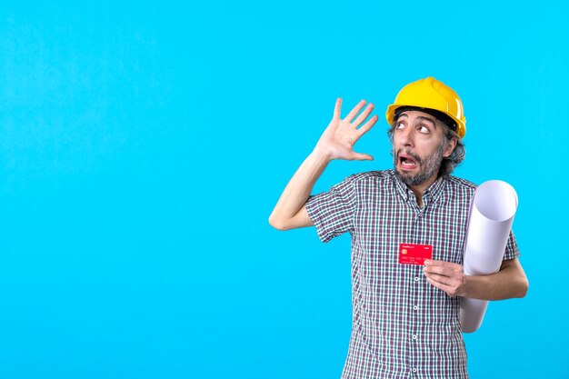 Vue de face constructeur masculin tenant un plan et une carte bancaire sur fond bleu constructeur de conception d'argent bâtiment travail travail architecture de couleur