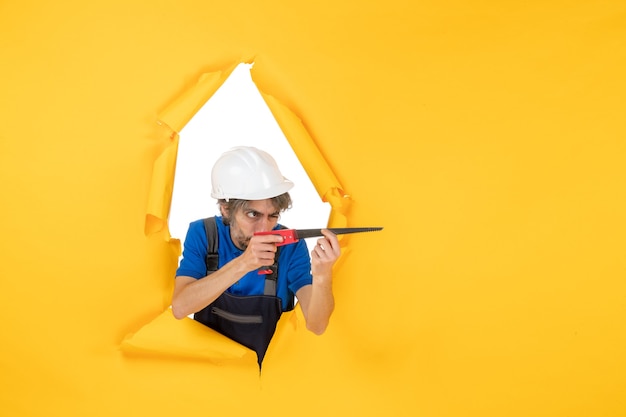 Vue de face constructeur masculin en uniforme avec instrument dans ses mains sur un bâtiment de couleur de travailleur de constructeur de travail de mur jaune