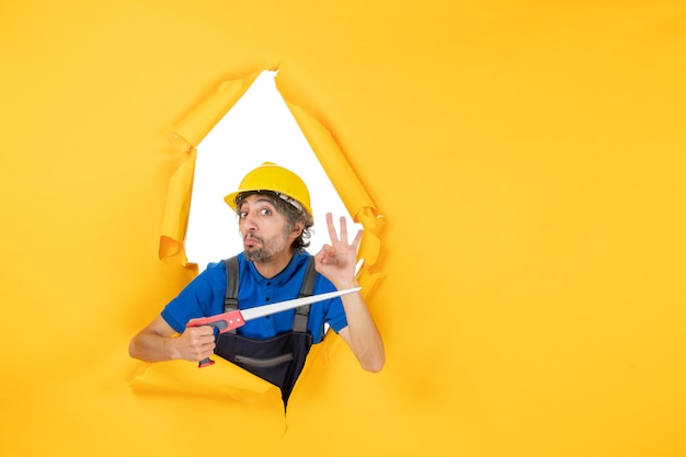 Vue de face constructeur masculin en uniforme avec instrument dans ses mains sur le travail du constructeur de travail de construction de mur jaune