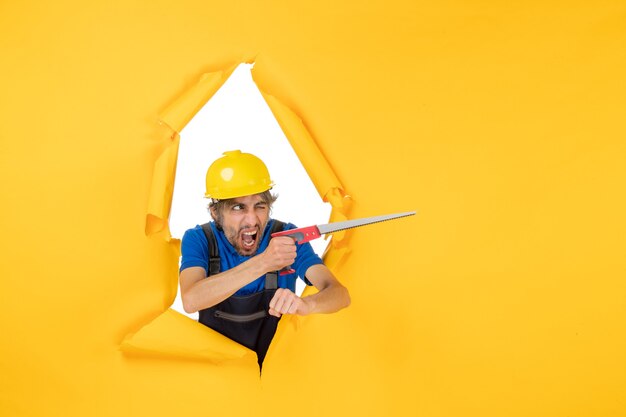 Vue de face constructeur masculin en uniforme avec instrument dans ses mains sur le travail de travailleur de travail de constructeur de couleur de bâtiment de mur jaune