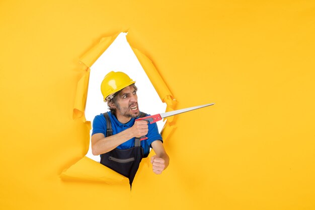 Vue de face constructeur masculin en uniforme avec instrument dans ses mains sur le travail de travailleur de travail de constructeur de couleur de bâtiment de mur jaune