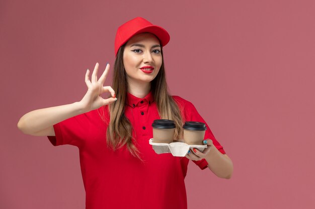 Vue de face de courrier féminin en uniforme rouge tenant des tasses de café de livraison brun souriant sur fond rose clair de livraison de services de travail uniforme de travail des femmes entreprise