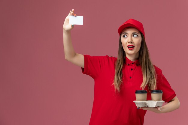 Vue de face de courrier féminin en uniforme rouge tenant des tasses de café de livraison et une carte sur l'uniforme de livraison d'emploi de fond rose