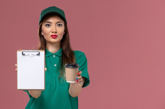 Vue de face de courrier féminin en uniforme vert et cape tenant la tasse de café de livraison et le bloc-notes sur le mur rose de travail de service de livraison uniforme entreprise féminine