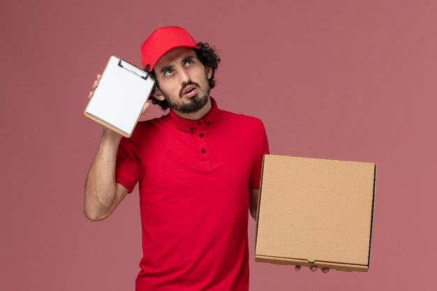 Vue de face de courrier masculin en chemise rouge et cape tenant la boîte de nourriture de livraison et le bloc-notes pensant sur le mur rose