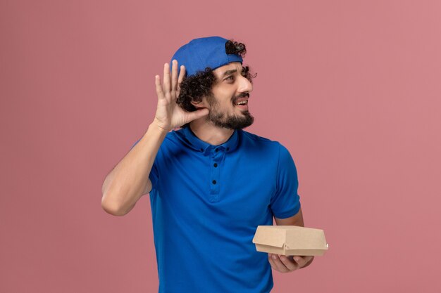 Vue de face de courrier masculin en uniforme bleu et cape tenant peu de colis de nourriture de livraison et en essayant d'entendre sur le mur rose des employés de l'entreprise de livraison uniforme