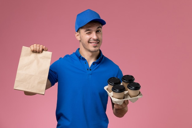 Vue de face de courrier masculin en uniforme bleu tenant des tasses de café de paquet de nourriture sur le mur rose, travailleur de livraison de service uniforme