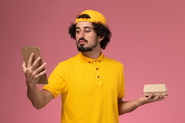 Vue de face de courrier masculin en uniforme jaune et cape avec bloc-notes et petit paquet de nourriture de livraison sur ses mains sur fond rose.