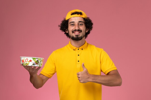 Vue de face de courrier masculin en uniforme jaune et cape avec bol de livraison rond sur ses mains sur le fond rose clair.