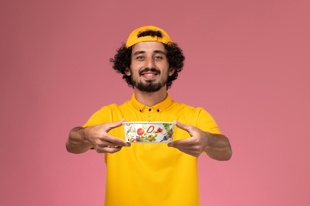 Vue de face de courrier masculin en uniforme jaune et cape avec bol de livraison rond sur ses mains sur le fond rose.