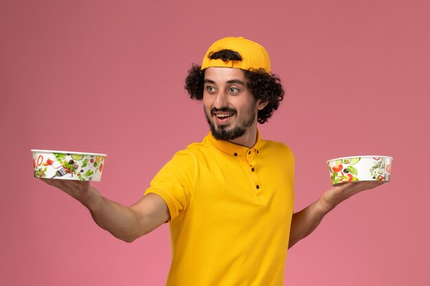 Vue de face de courrier masculin en uniforme jaune et cape avec des bols de livraison ronds sur ses mains sur le fond rose clair.