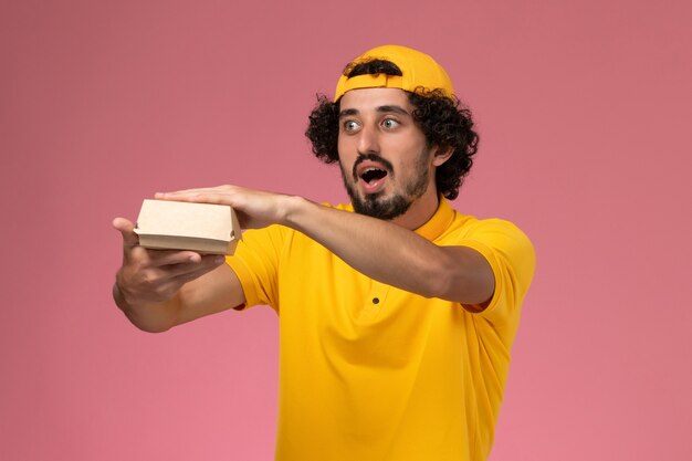Vue de face de courrier masculin en uniforme jaune et cape avec petit paquet de nourriture de livraison sur ses mains sur fond rose clair.