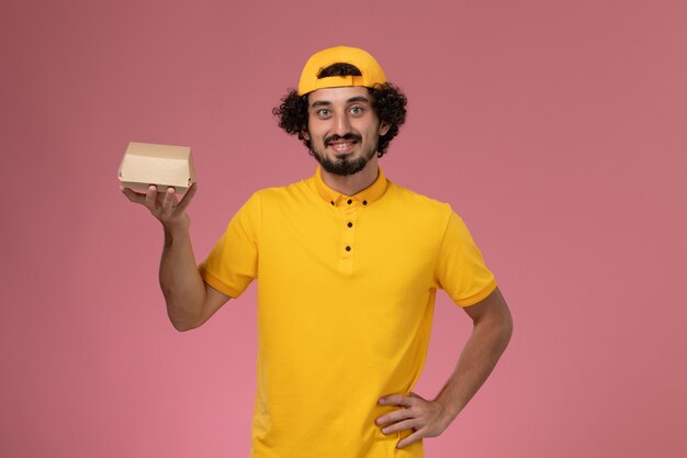 Vue de face de courrier masculin en uniforme jaune et cape avec petit paquet de nourriture de livraison sur ses mains sur le fond rose.