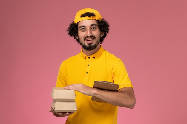 Vue de face de courrier masculin en uniforme jaune et cape avec de petits paquets de nourriture de livraison et bloc-notes sur ses mains sur le fond rose clair.