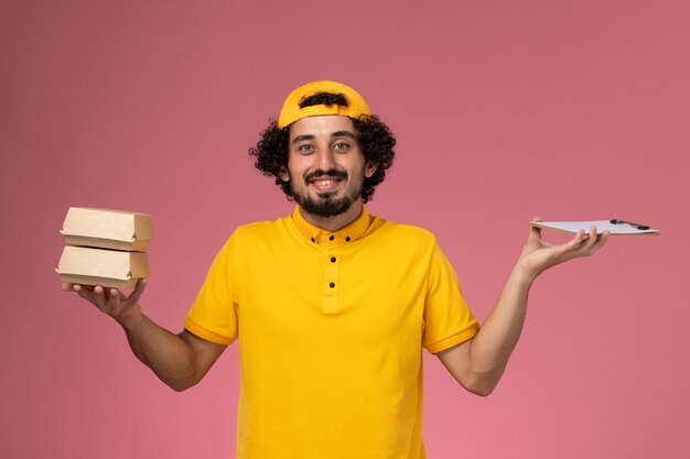 Vue de face de courrier masculin en uniforme jaune et cape avec de petits paquets de nourriture de livraison et bloc-notes sur ses mains sur le fond rose clair.