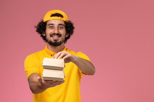 Vue de face de courrier masculin en uniforme jaune et cape avec peu de colis alimentaires de livraison sur ses mains sur le fond rose.