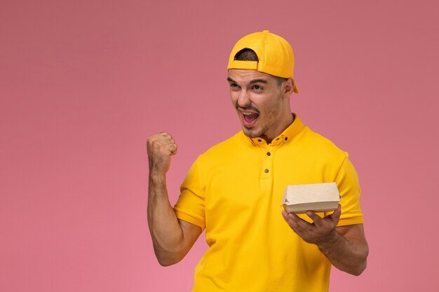 Vue de face de courrier masculin en uniforme jaune et cape tenant petit paquet de nourriture de livraison et acclamant sur le fond rose.