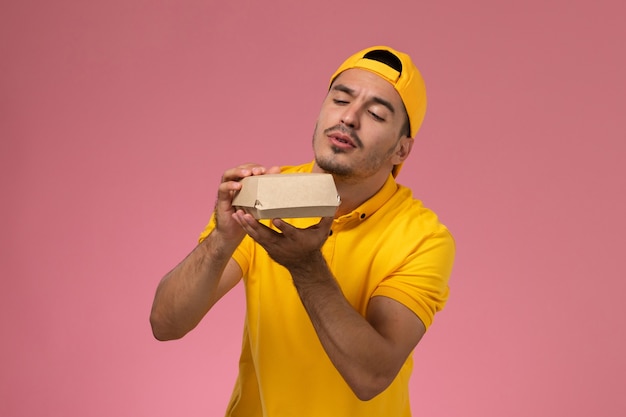 Vue de face de courrier masculin en uniforme jaune et cape tenant petit paquet de nourriture de livraison sur fond rose clair.