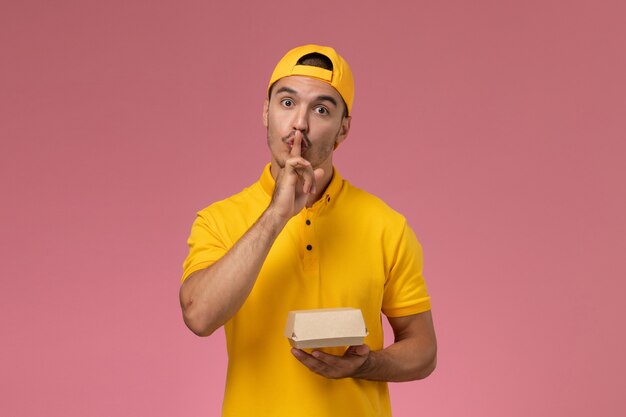 Vue de face de courrier masculin en uniforme jaune et cape tenant petit paquet de nourriture de livraison sur fond rose.