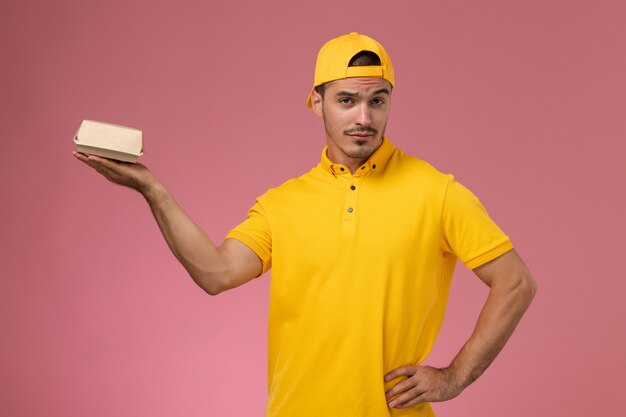 Vue de face de courrier masculin en uniforme jaune et cape tenant petit paquet de nourriture de livraison sur fond rose.