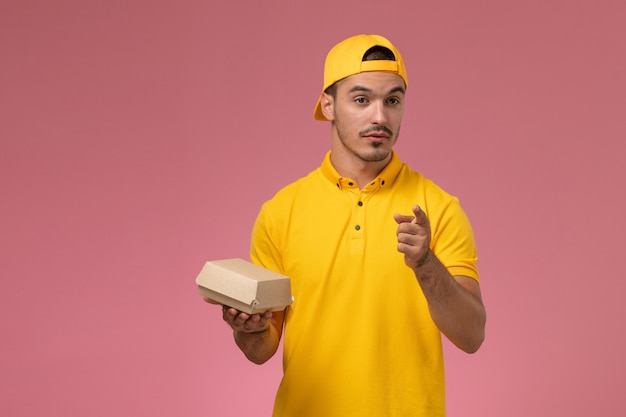 Vue de face de courrier masculin en uniforme jaune et cape tenant petit paquet de nourriture de livraison et parler sur fond rose clair.