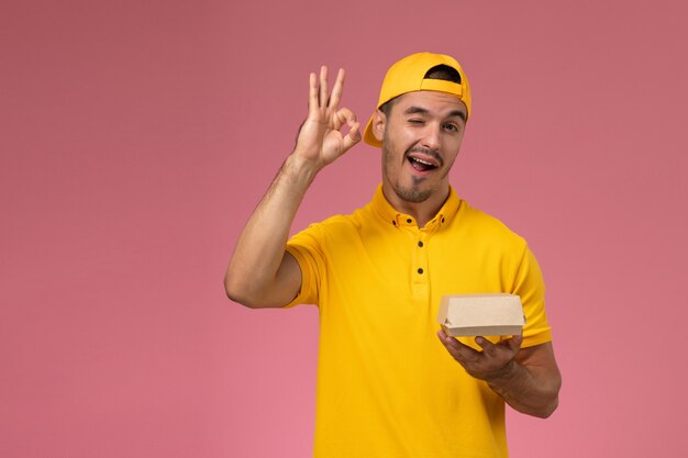 Vue de face de courrier masculin en uniforme jaune et cape tenant peu de colis de livraison de nourriture clignotant sur fond rose.