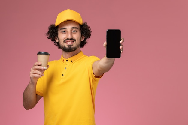 Vue de face de courrier masculin en uniforme jaune et cape tenant la tasse de café de livraison et téléphone sur mur rose