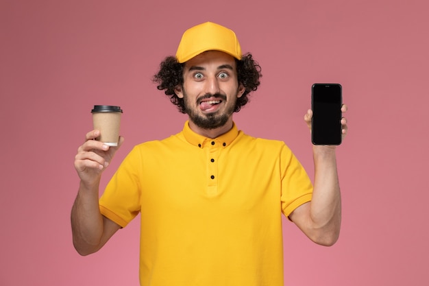 Vue de face de courrier masculin en uniforme jaune et cape tenant la tasse de café de livraison et téléphone sur mur rose
