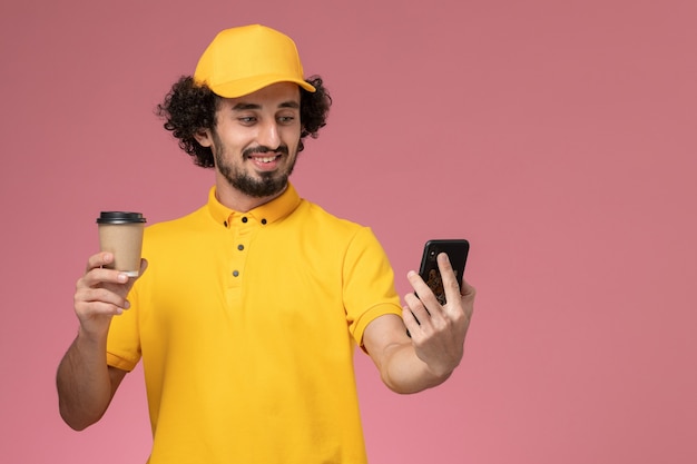 Vue de face de courrier masculin en uniforme jaune et cape tenant la tasse de café de livraison et téléphone sur mur rose