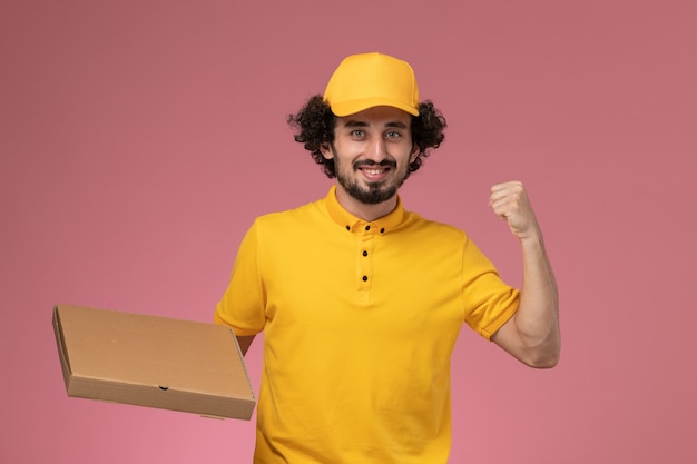 Vue de face de courrier masculin en uniforme jaune tenant la boîte de livraison de nourriture applaudissant sur le mur rose clair