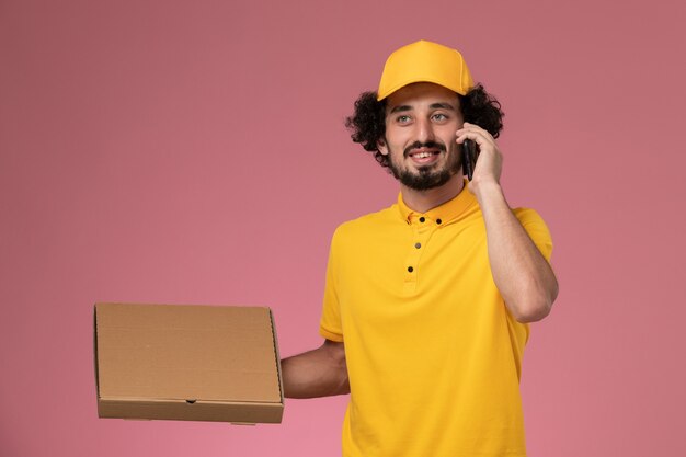 Vue de face de courrier masculin en uniforme jaune tenant la boîte de livraison de nourriture et parler au téléphone sur le mur rose clair