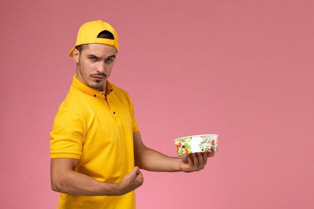 Vue de face de courrier masculin en uniforme jaune tenant le bol de livraison et posant sur fond rose.