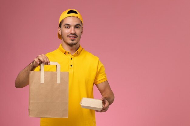 Vue de face de courrier masculin en uniforme jaune tenant des colis alimentaires de livraison sur le fond rose clair.