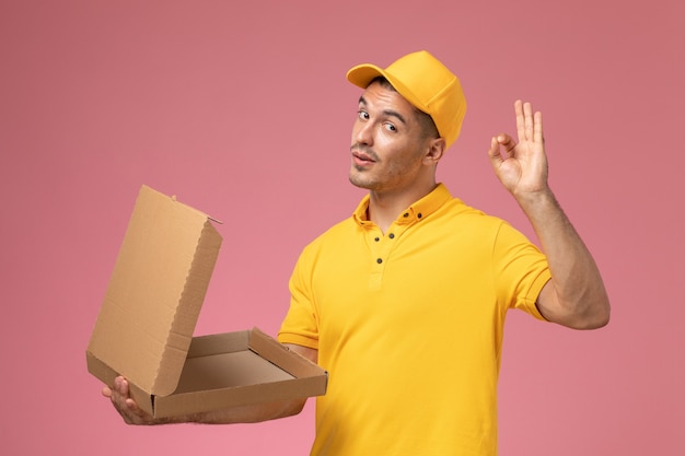 Vue de face de courrier masculin en uniforme jaune tenant et ouvrant la boîte de livraison de nourriture vide sur le bureau rose