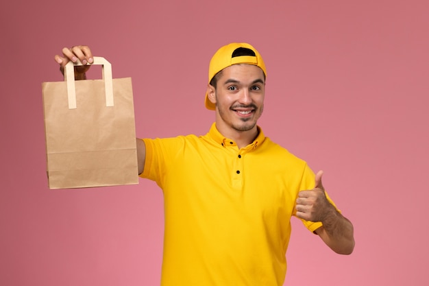 Vue de face de courrier masculin en uniforme jaune tenant le paquet de papier de livraison souriant sur fond rose clair.