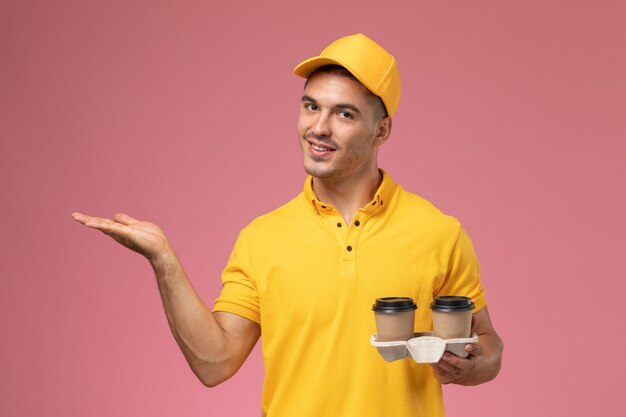 Vue de face de courrier masculin en uniforme jaune tenant des tasses de café de livraison souriant sur un bureau rose clair