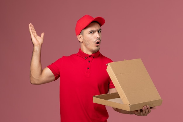 Vue de face de courrier masculin en uniforme rouge et cape tenant la boîte de nourriture avec un sourire sur le mur rose de travail de service de l'homme uniforme de livraison