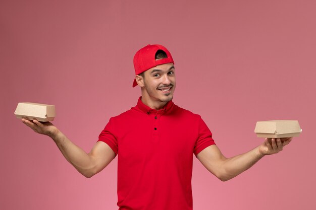 Vue de face de courrier masculin en uniforme rouge et cape tenant de petits colis de livraison sur le fond rose.