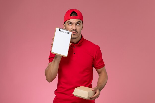 Vue de face de courrier masculin en uniforme rouge et cape tenant peu de colis de livraison avec bloc-notes sur fond rose.