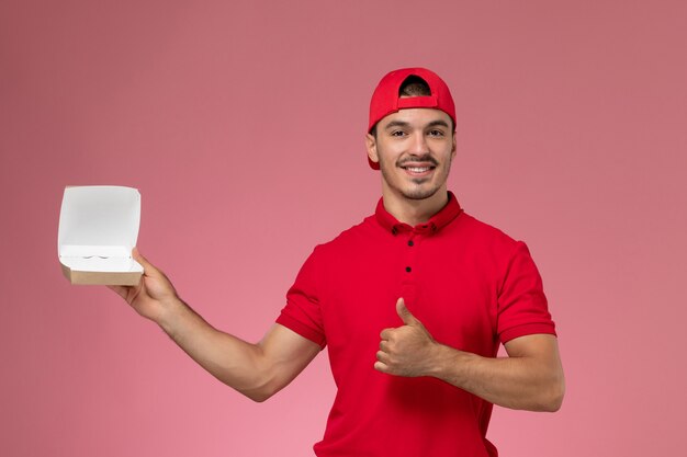 Vue de face de courrier masculin en uniforme rouge et cape tenant peu de colis de livraison souriant sur le fond rose.