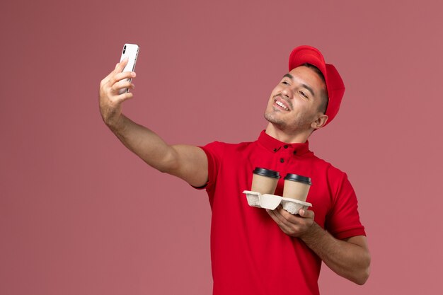 Vue de face de courrier masculin en uniforme rouge tenant des tasses de café de livraison brun prenant un selfie avec eux sur le mur rose