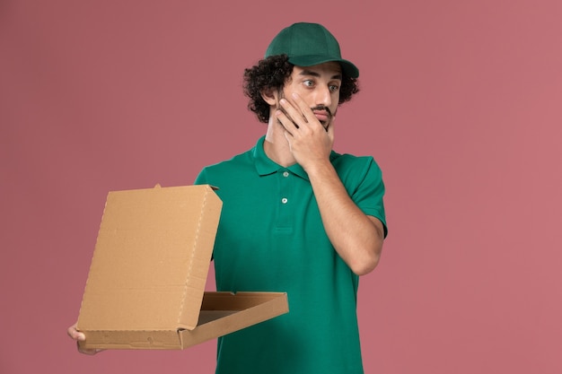 Vue de face de courrier masculin en uniforme vert et cape tenant la boîte de nourriture de livraison pensée sur fond rose service de livraison uniforme